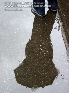 Picture of puddle with a reflected silhouette of a person on a street corner.  The person is holding an umbrella and the raindrops are visible in the puddle.  www.FreePhotoCourse.com