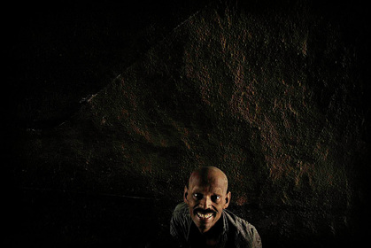 Picture of an Indian man sitting in the darkness of an impoverished existance, a mud wall behind him.  Despite the conditions, he wears a smile and his eyes are bright and happy.Part of the Photographer Profiles series at FreePhotoCourse.com. © 2010, Soham Gupta, all rights reserved.      